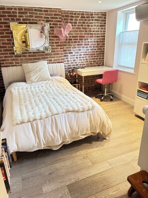 A bedroom with a brick wall, a bed with a white duvet, a desk with a pink chair, and a window with blinds. - NEW! Gracious Family Townhome 18 Minutes to Manhattan (Jersey City)