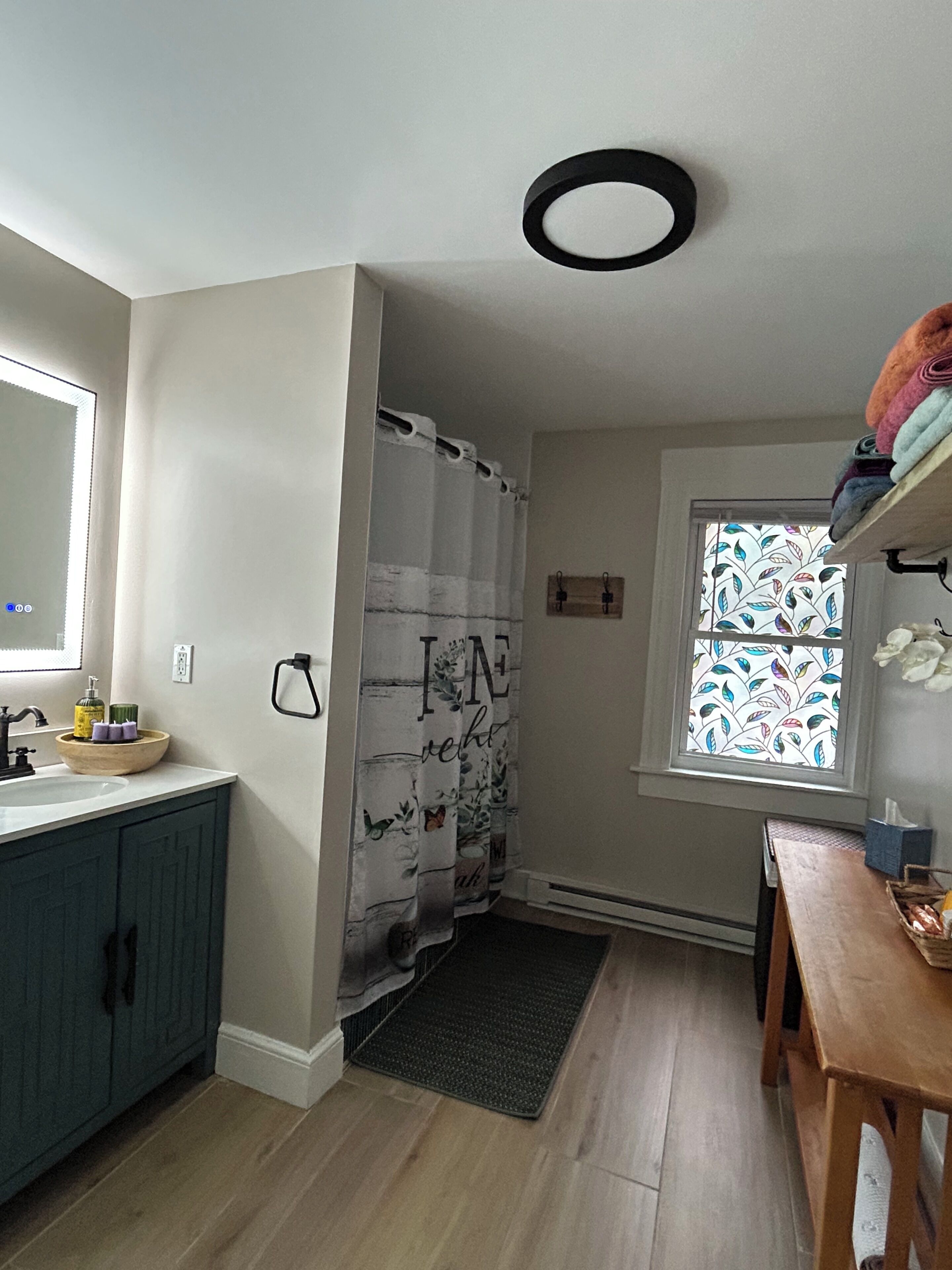 Een badkamer met douchegordijn, wastafel, spiegel, raam met bladmotief en een houten plank met handdoeken.