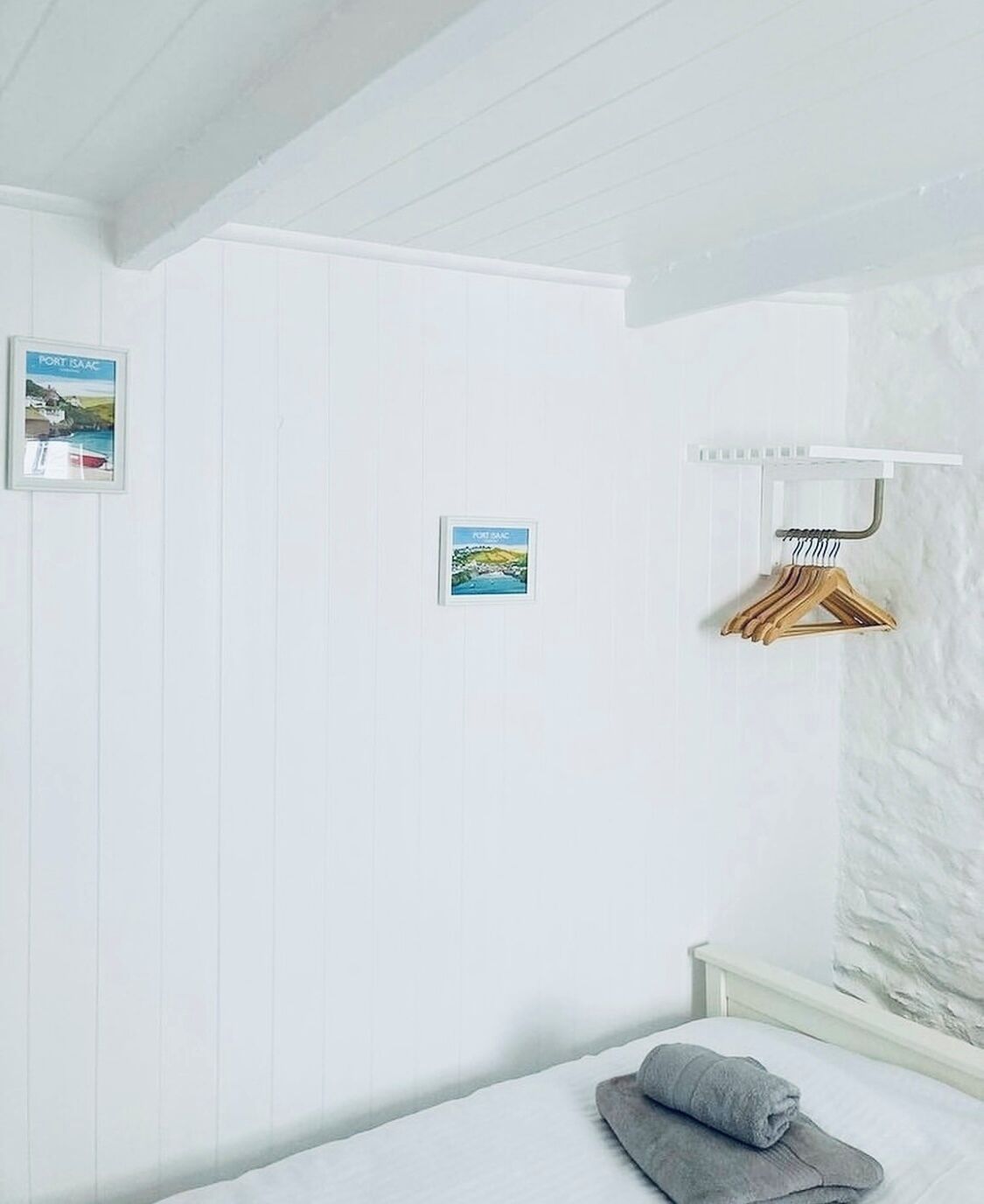 Une pièce blanche avec un lit, une serviette, un porte-manteau en bois et deux tableaux encadrés accrochés au mur.