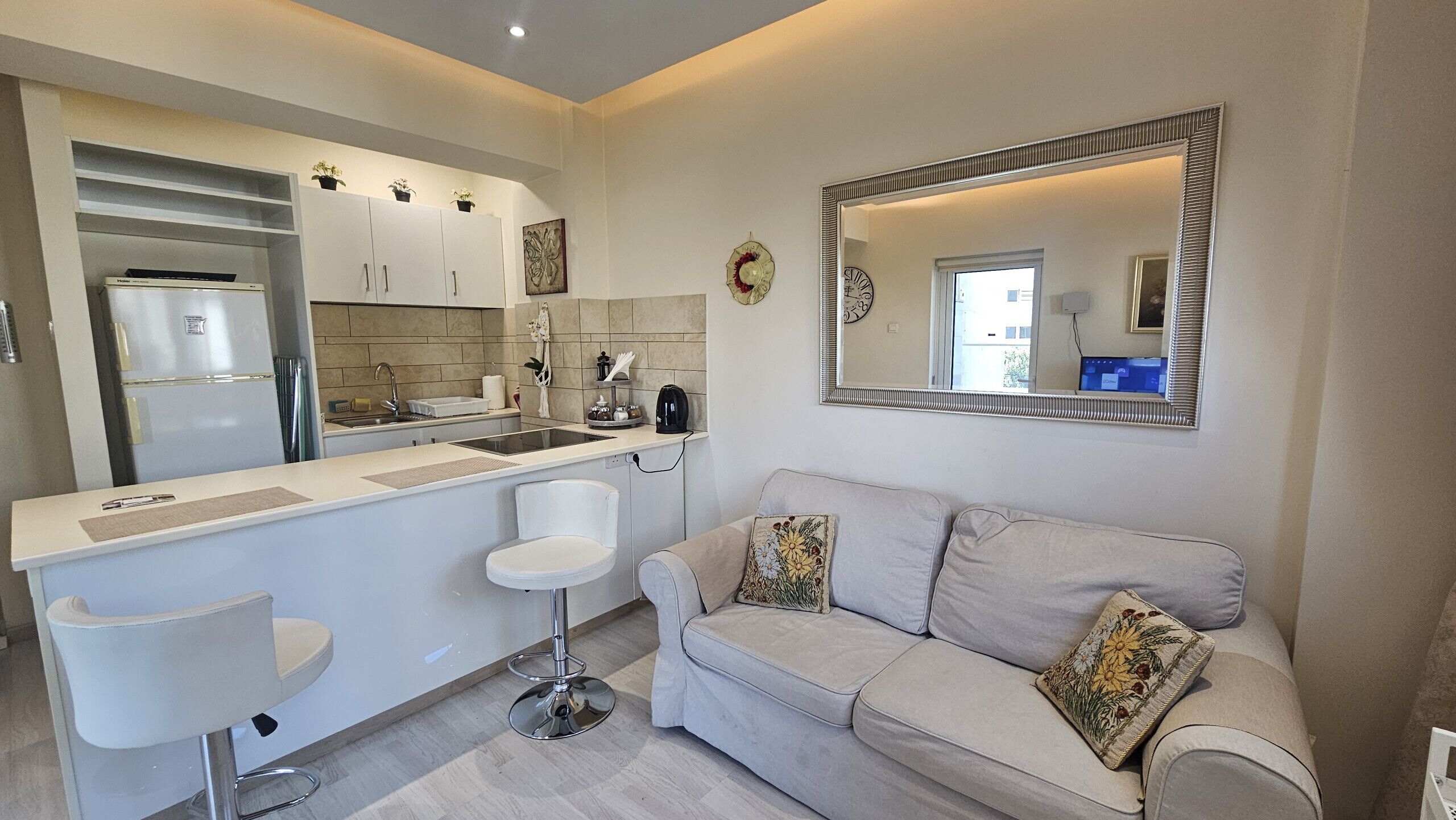 A modern kitchen with white cabinets, a built-in oven, and a sink. There is a white bar stool and a beige sofa with floral cushions. A large mirror with a decorative frame hangs on the wall.