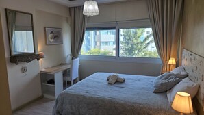 Quarto de hotel com cama, escrivaninha, cadeira, abajur, espelho e janela com cortinas.