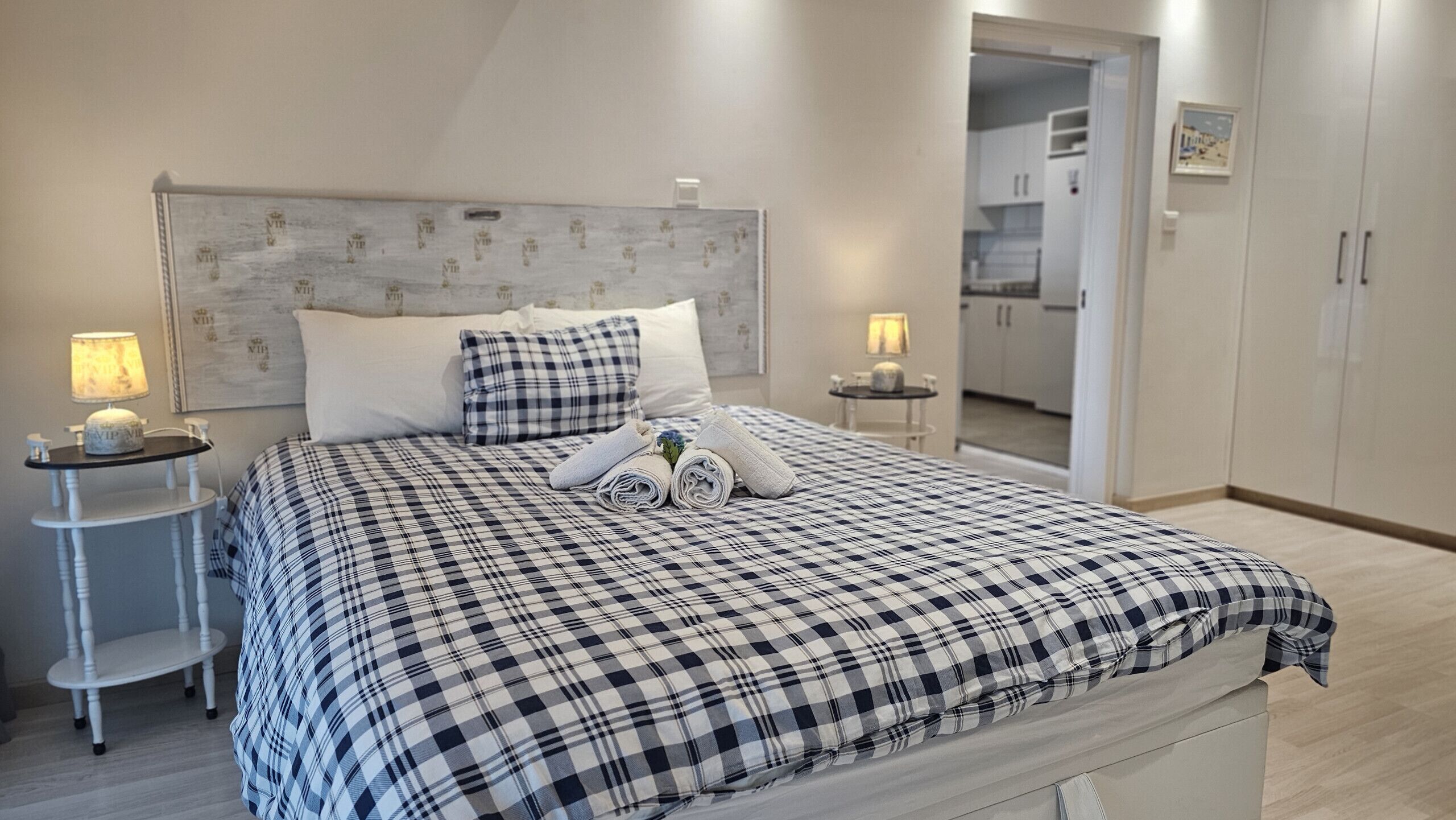 A bedroom with a checkered bedspread, two bedside tables with lamps, a headboard, and a view into the kitchen.