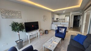 A modern living room with a flat-screen TV, a coffee table, and seating. A kitchen is visible in the background. - 116 NIC 302b - Stay Armonia Central Suite (Nicosia)
