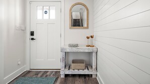 Une porte blanche avec une petite fenêtre, une console en bois rustique surmontée d’un miroir et, en dessous, un panier en osier.