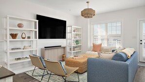 Un salon avec un téléviseur, des étagères blanches, un canapé bleu, des fauteuils en osier et un tapis à motifs.