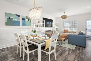 A dining area with a wooden table, white chairs, and a chandelier. There is a sofa and a TV in the background. - Modern and sophisticated beach retreat, two blocks from beach & boardwalk. (Ocean City)