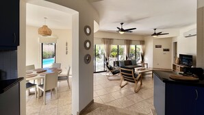 Amplia sala de estar con mesa de comedor, sillas y sofá. Cuenta con ventiladores de techo, televisor y una zona de cocina con armarios y encimera.