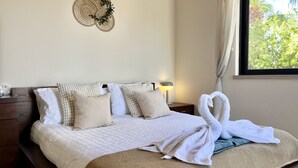 Una cama bien hecha con una colcha blanca y beige, dos almohadas, un tapiz decorativo, una mesita de noche con una lámpara y una ventana con cortinas.