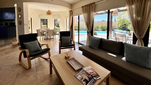 Amplia sala de estar con sofá, dos sillones, una mesa de centro y una mesa de comedor. Se observa un ventilador de techo y ventanas grandes con cortinas.