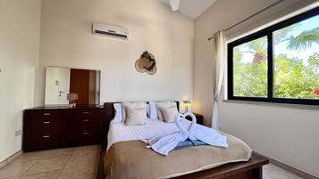 A bedroom with a bed, a ceiling fan, a wall-mounted air conditioner, a bedside lamp, a window with a view of greenery, and a wooden nightstand.