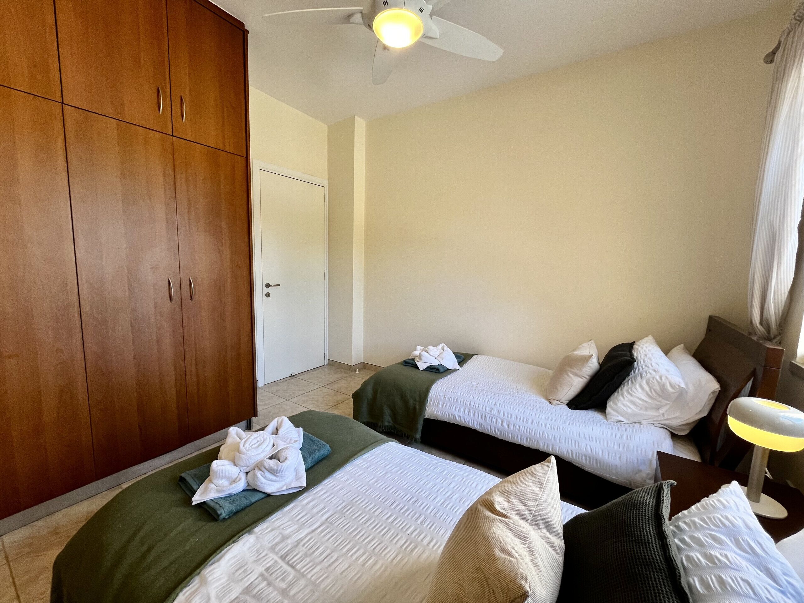 Habitación de hotel con dos camas, un armario de madera, un ventilador de techo y una mesita de noche con lámpara.