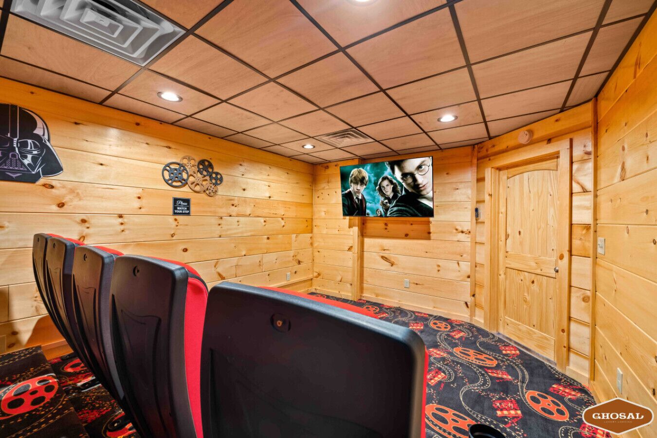 Ein gemütlicher Raum mit Holzwänden, einem großen Fernsehbildschirm, der eine Filmszene zeigt, einer bequemen Sitzecke mit roten und schwarzen Stühlen und einem Schild an der Wand mit der Aufschrift 'Vorsicht, ein stolpergefährdeter Bereich'.