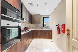 A modern kitchen with a built-in oven, a stainless steel range hood, and a marble countertop. There are wooden cabinets and a window providing natural light. - StayShort - Hidden Jewel 1 minute walk from Al Furjan Metro Sleeps 5 (Dubai)