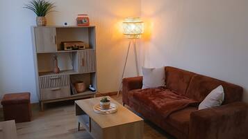 Een moderne woonkamer met een bruine bank, een houten salontafel en een vloerlamp. Er is een boekenkast met decoratieve objecten en een klein tafeltje met een plant.