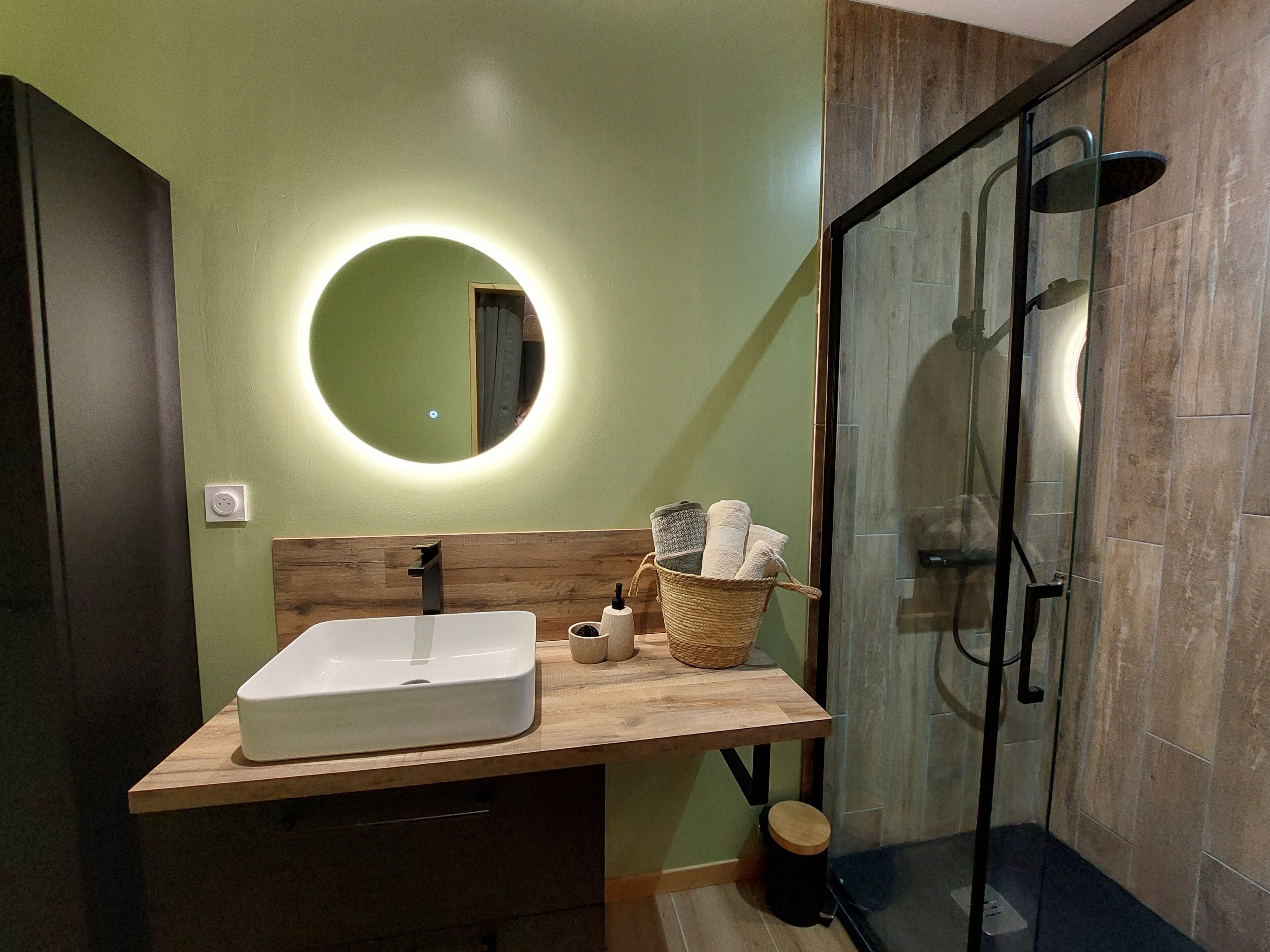 Une salle de bain moderne avec un meuble-lavabo en bois, un lavabo blanc, un miroir rond et une cabine de douche en verre.