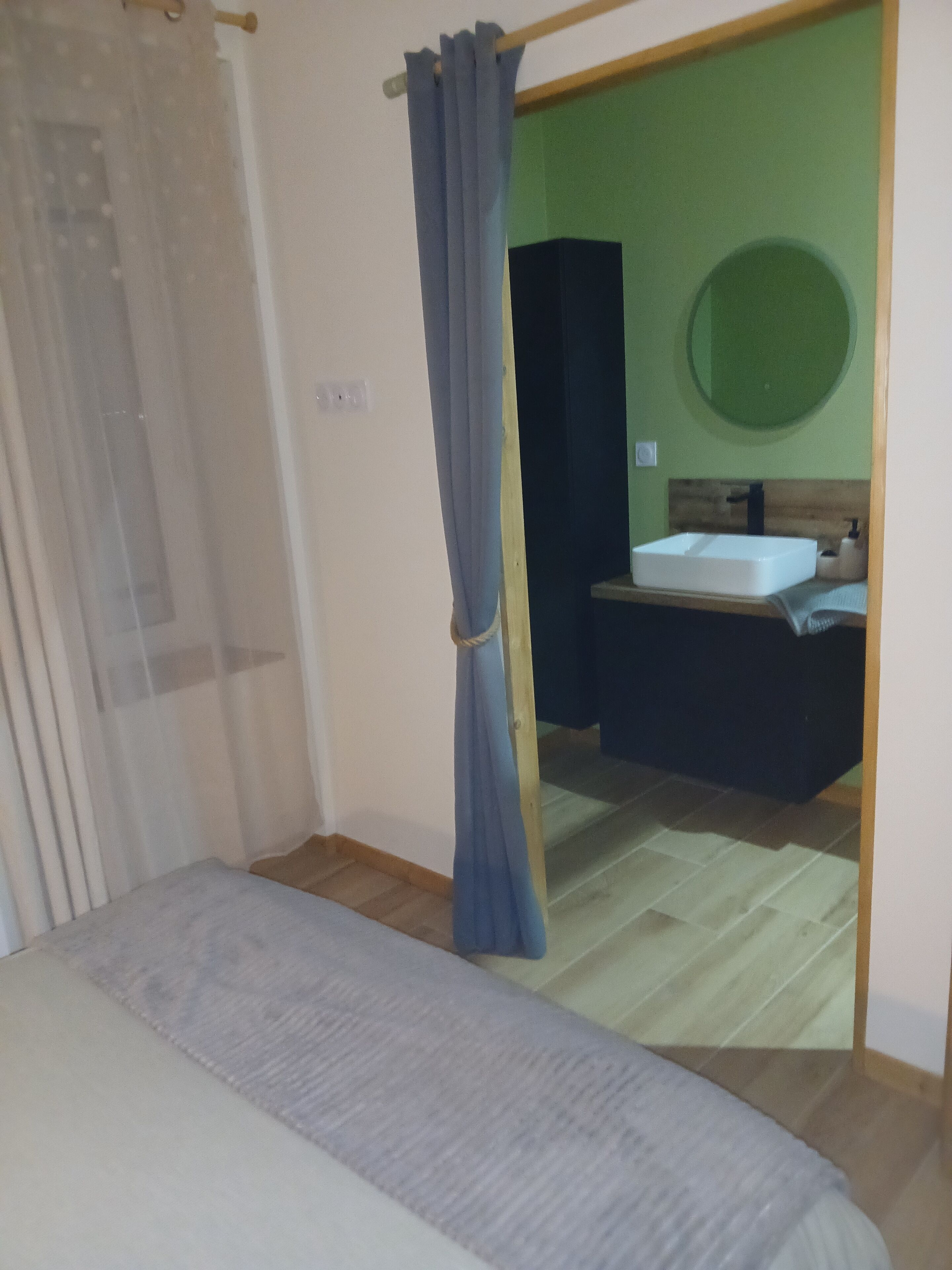 Ein Zimmer mit einem Bett, einem hölzernen Kopfteil, einer grünen Wand, einem runden Spiegel und einem Badezimmer mit Waschbecken und Handtuchhalter.