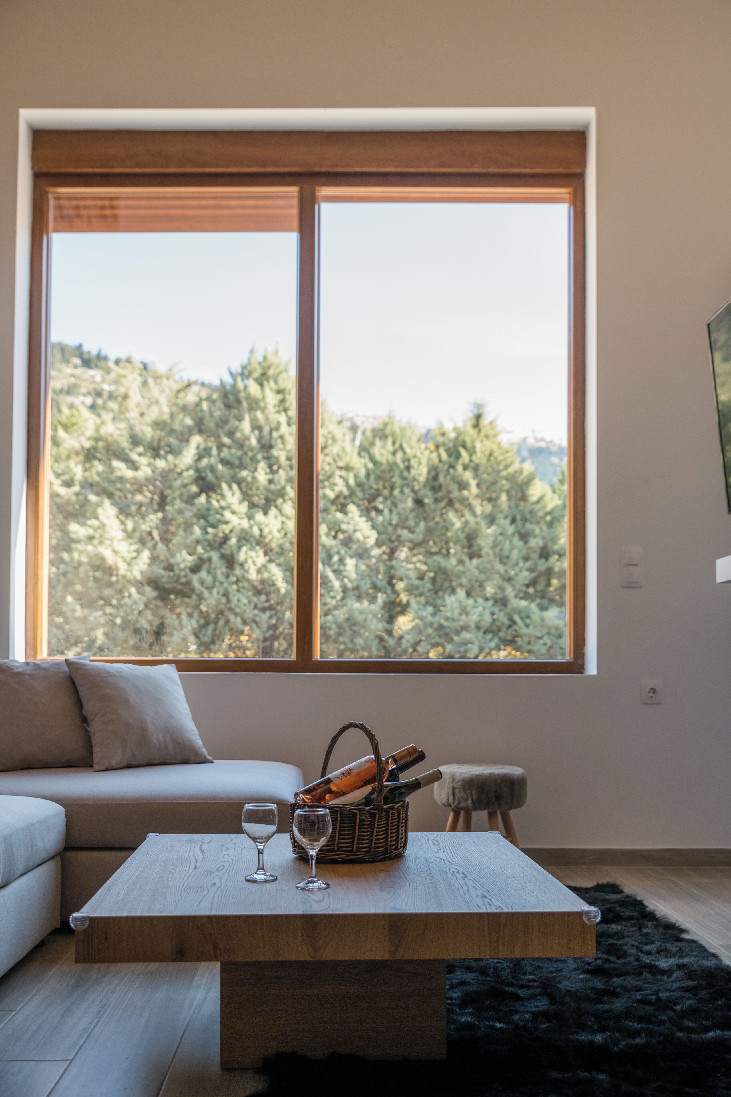 A room with a large window, a wooden coffee table, a basket, two wine glasses, a sofa, and a small stool.
