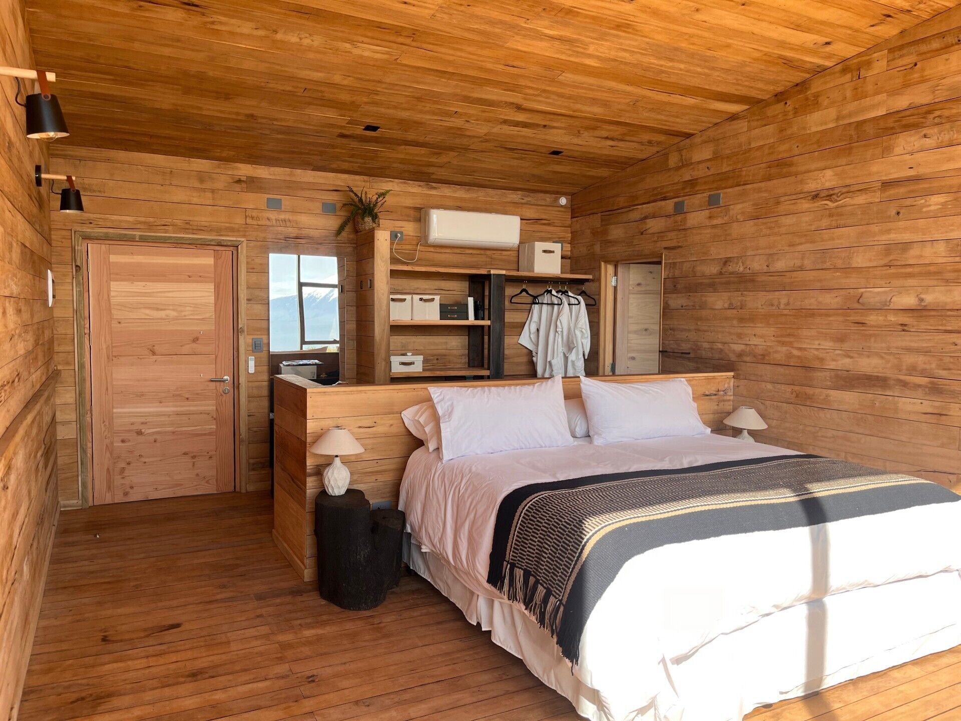 Un dormitorio de cabaña de madera con una cama, lámparas de noche, un aparador de madera y una ventana con vistas.