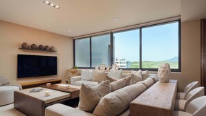 A modern living room with a large flat-screen TV, a sofa set, a coffee table, and a view of a mountainous landscape through large windows. - Patamar Luxury Condo - Includes Chef, Maid & Premier Access (Punta Mita)