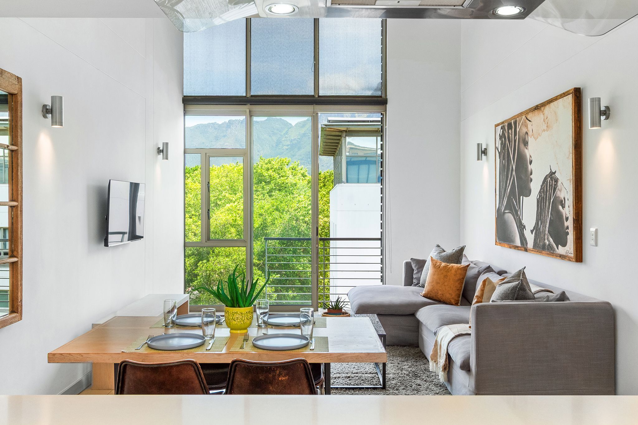 Un comedor moderno con una mesa de madera dispuesta para cuatro personas, un sofá con cojines, y un ventanal que ofrece vistas a zonas verdes y montañas.