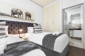 A hotel room with two beds, a large tufted headboard, a nightstand with a lamp, a painting of elephants, a wardrobe, and a bathroom with a sink and mirror. - The Quadrant 406 (Cape Town)