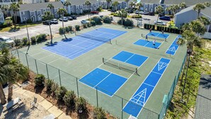 Un court de tennis entouré d’une clôture, avec vue sur l’océan et des immeubles d’habitation en arrière-plan.