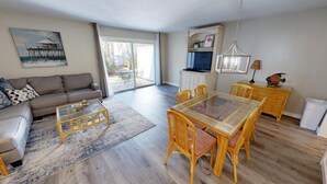 Un vaste espace de vie comprenant un canapé, une table basse et une table à manger avec des chaises. Un téléviseur est fixé au mur, et il y a une fenêtre avec des rideaux.