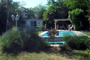 Una piscina rodeada de abundante vegetación, un pabellón y un pequeño edificio con un campanario.