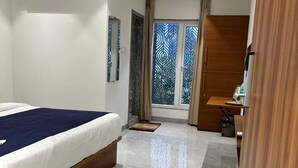 A hotel room with a bed, a wooden wardrobe, a desk, and a window with decorative curtains. - HOTEL MANGALAM REGENCY (Indore)