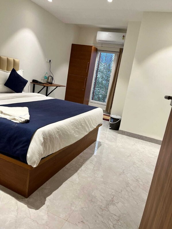 A hotel room with a bed, a bedside table with a lamp, a window with a decorative view, and an air conditioning unit.
