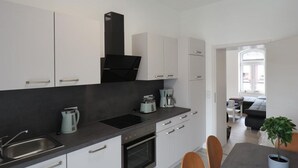 A modern kitchen with white cabinets, a black range hood, and a built-in oven. There is a dining area with a table and chairs, and a potted plant on the table.