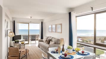 A modern living room with a balcony overlooking the sea, a dining area with a table set for two, and a sofa with a view.