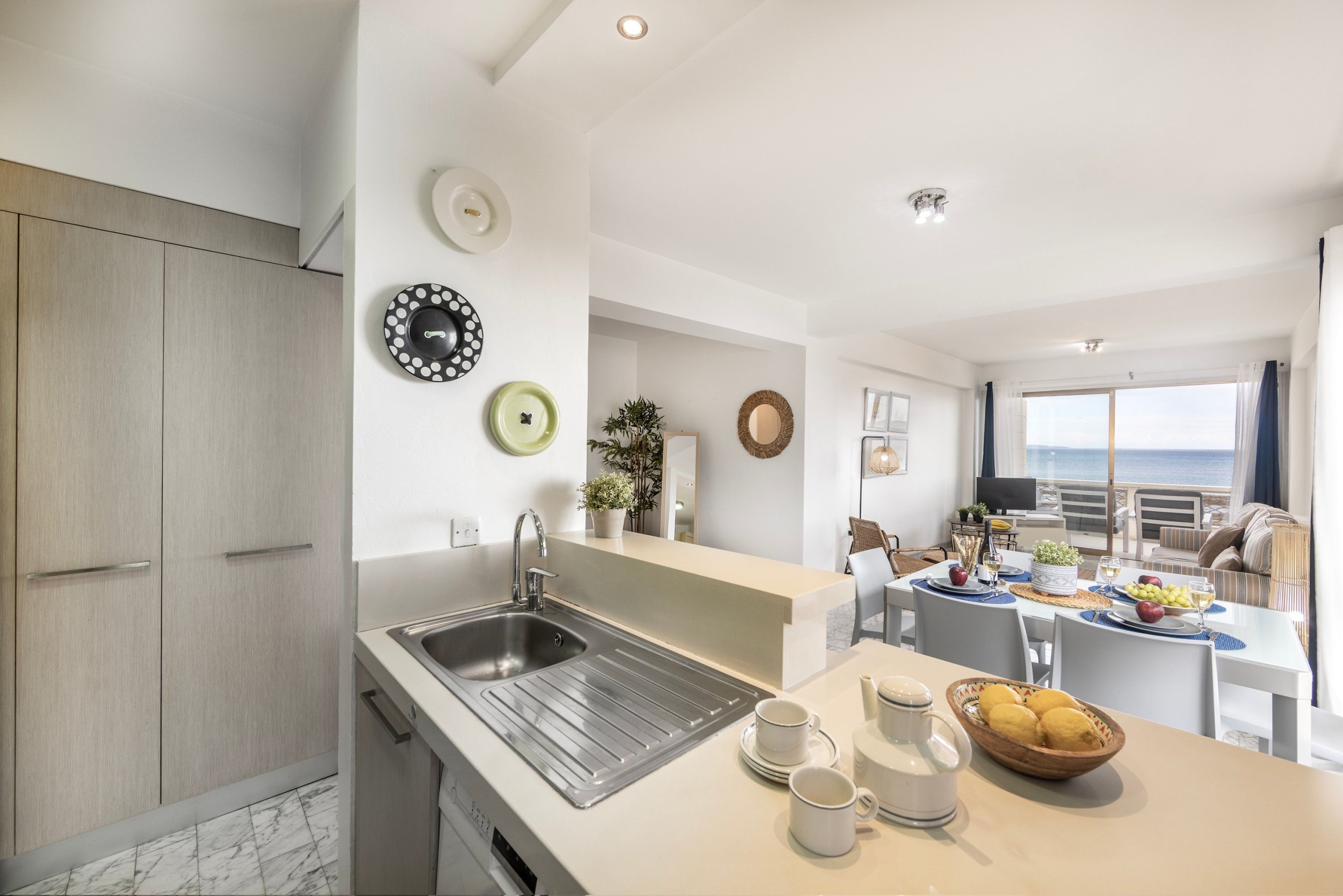A modern kitchen with a sink, dining area with a table set for two, and a view of the sea.