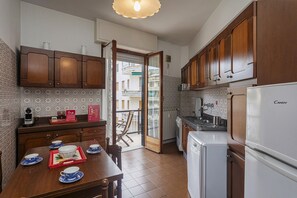 Eine kleine Küche mit Holzschränken, einem Esstisch, der mit blau-weißem Geschirr gedeckt ist, einem Kühlschrank und einem Fenster mit Blick auf einen Balkon.