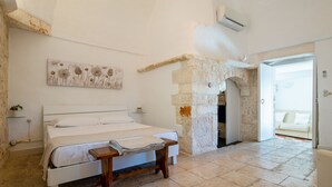 A bedroom with a bed, a wooden bench, a stone wall, and an open doorway leading to another room. - Campanile Apartment With Terrace by Wonderful Italy (Ostuni)