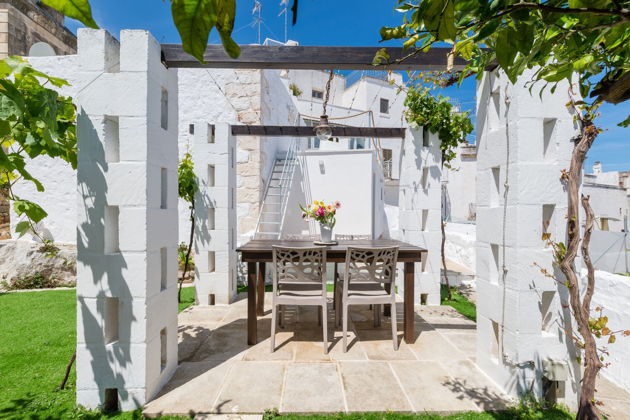 A white-walled courtyard with a table and chairs, a hanging light fixture, and a staircase leading to an upper level.