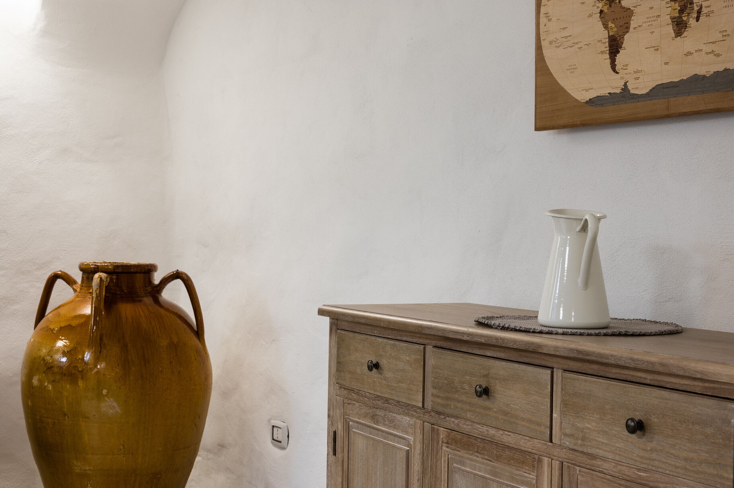 A room with a large brown vase on a wooden dresser, a white vase on a coaster, and a world map on the wall.