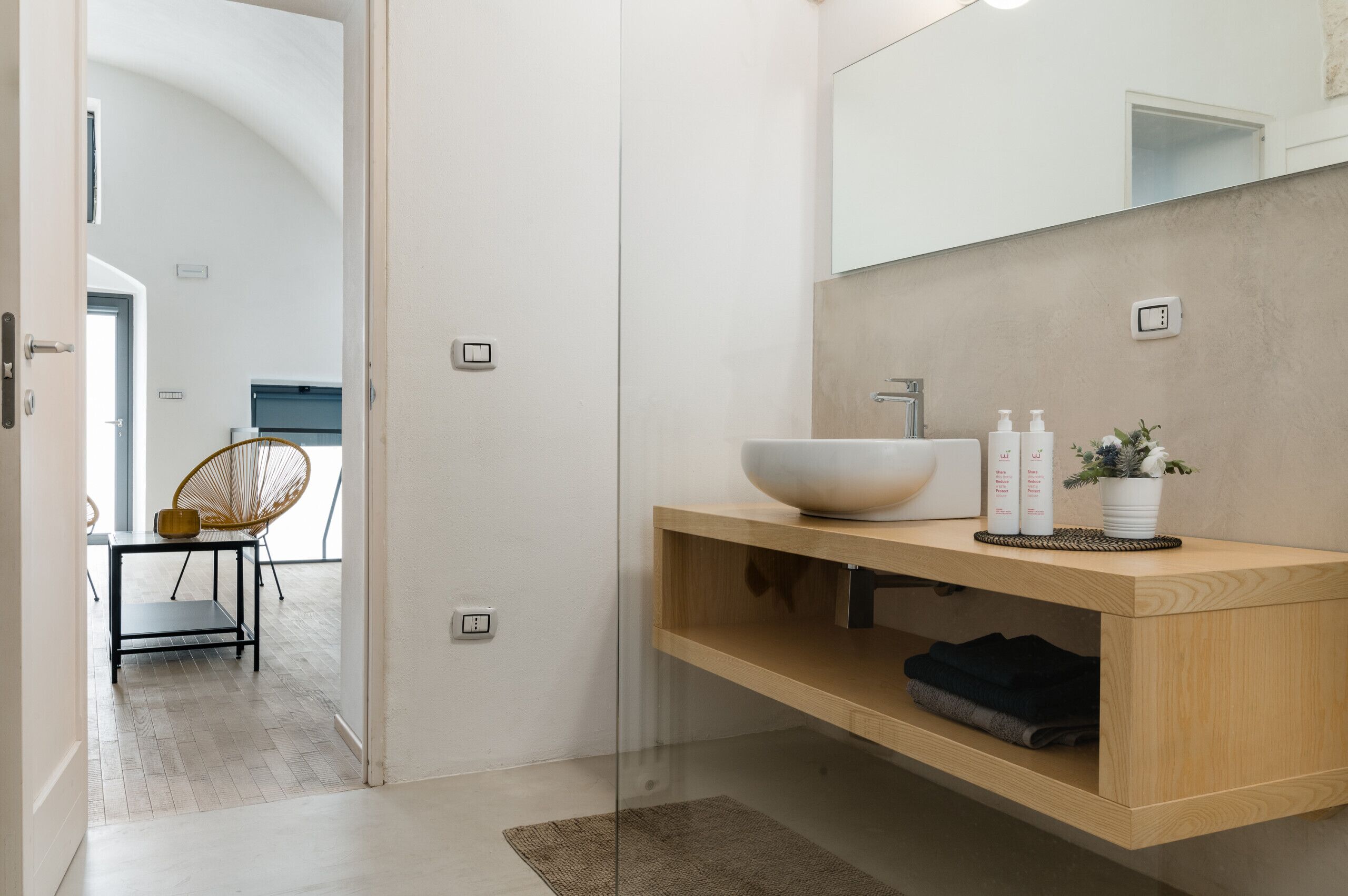 A modern bathroom with a large mirror, a wooden vanity, a bowl sink, and a chair in the adjacent room.