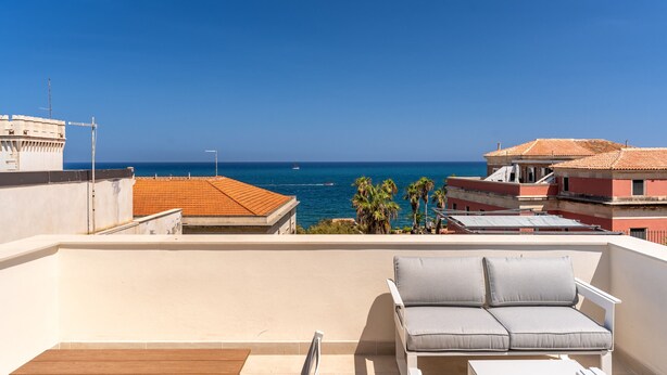 A rooftop terrace with a white sofa and a wooden table, overlooking a coastal view with buildings and palm trees. - Aquamarine Terrace by Wonderful Italy (Syracuse)