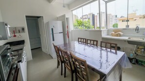 Een keuken met een grote eettafel, stoelen, een koelkast en een raam met uitzicht op gebouwen.