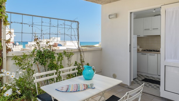 Ein Balkon mit Tisch, einem blauen Krug und einem Stuhl. Im Hintergrund sind Meer und Gebäude zu sehen.