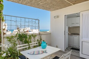Een balkon met een tafel, een blauwe kruik en een stoel. In de verte is de zee en gebouwen te zien.
