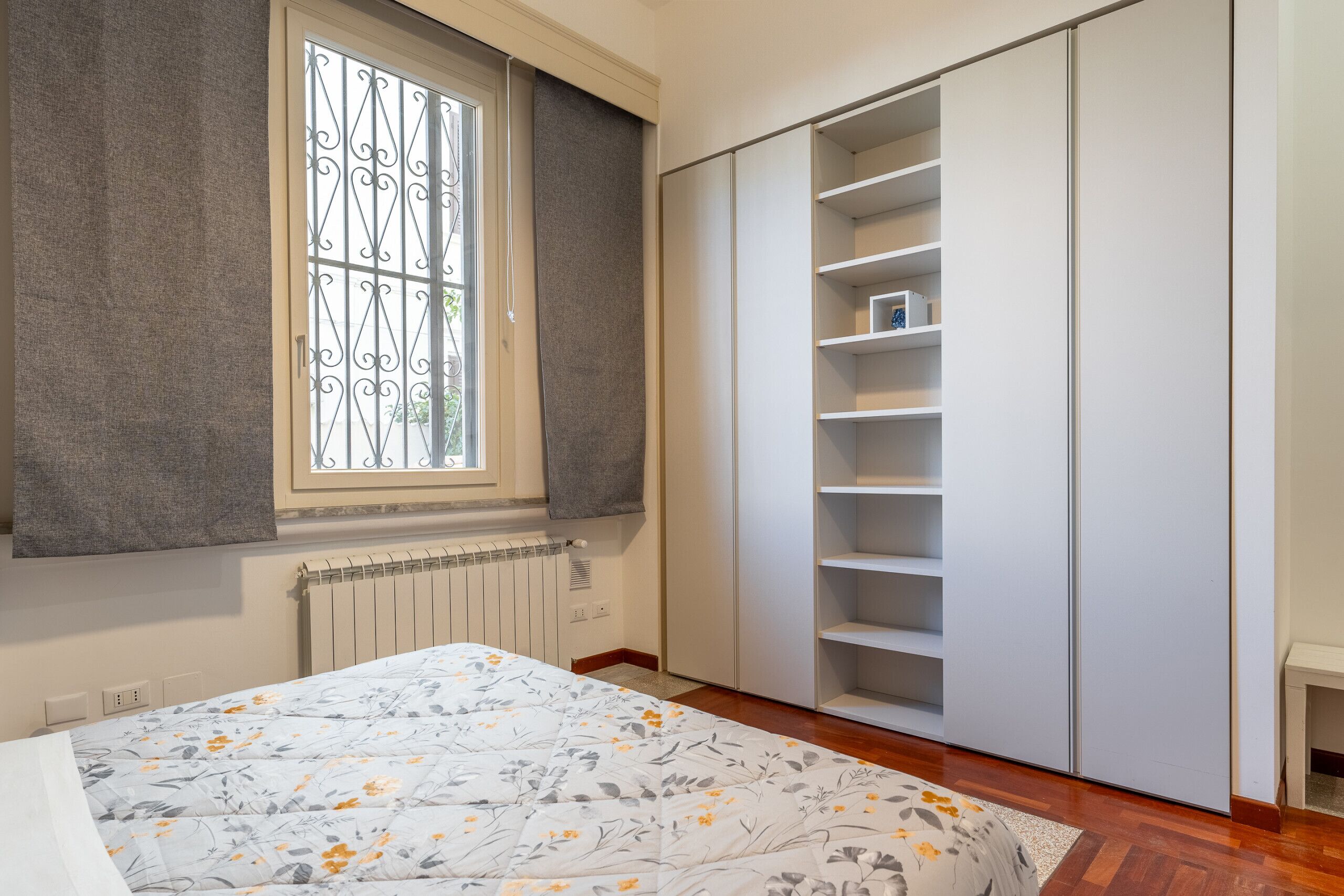A bedroom with a bed, a window with a curtain, a radiator, and a wardrobe with shelves.