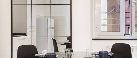 A modern dining area with a glass table, black chairs, and a view of a cityscape through a window.