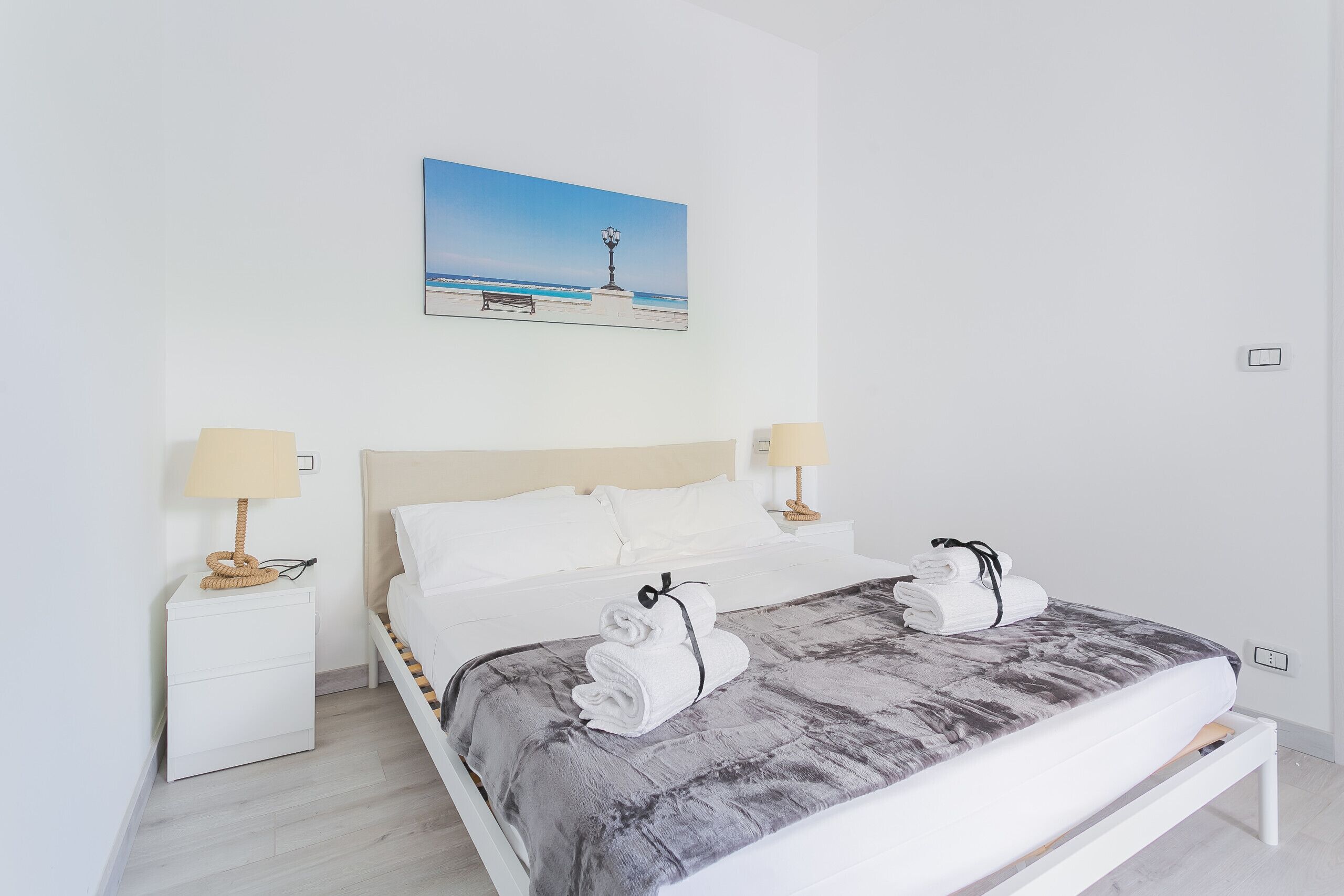 Habitación de hotel ordenada con una cama, dos mesitas de noche con lámparas, un cuadro de una playa y un banco.