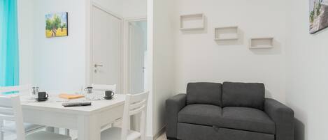 A modern living room with a grey sofa, white dining table, and chairs. There are three wall-mounted shelves and a door leading to another room.