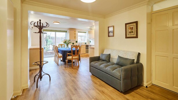 A spacious living area with a grey sofa, wooden dining table, and chairs, leading to a kitchen with wooden cabinets and a window. - Host & Stay | Riders Rest (Wootton)
