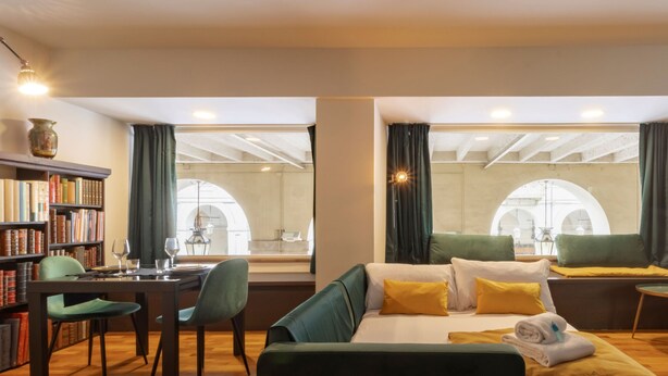 A hotel room with a bed, a sofa, a dining area with a table and chairs, a bookshelf, and large windows with curtains. - Turin Bed Books by Wonderful Italy (Turin)