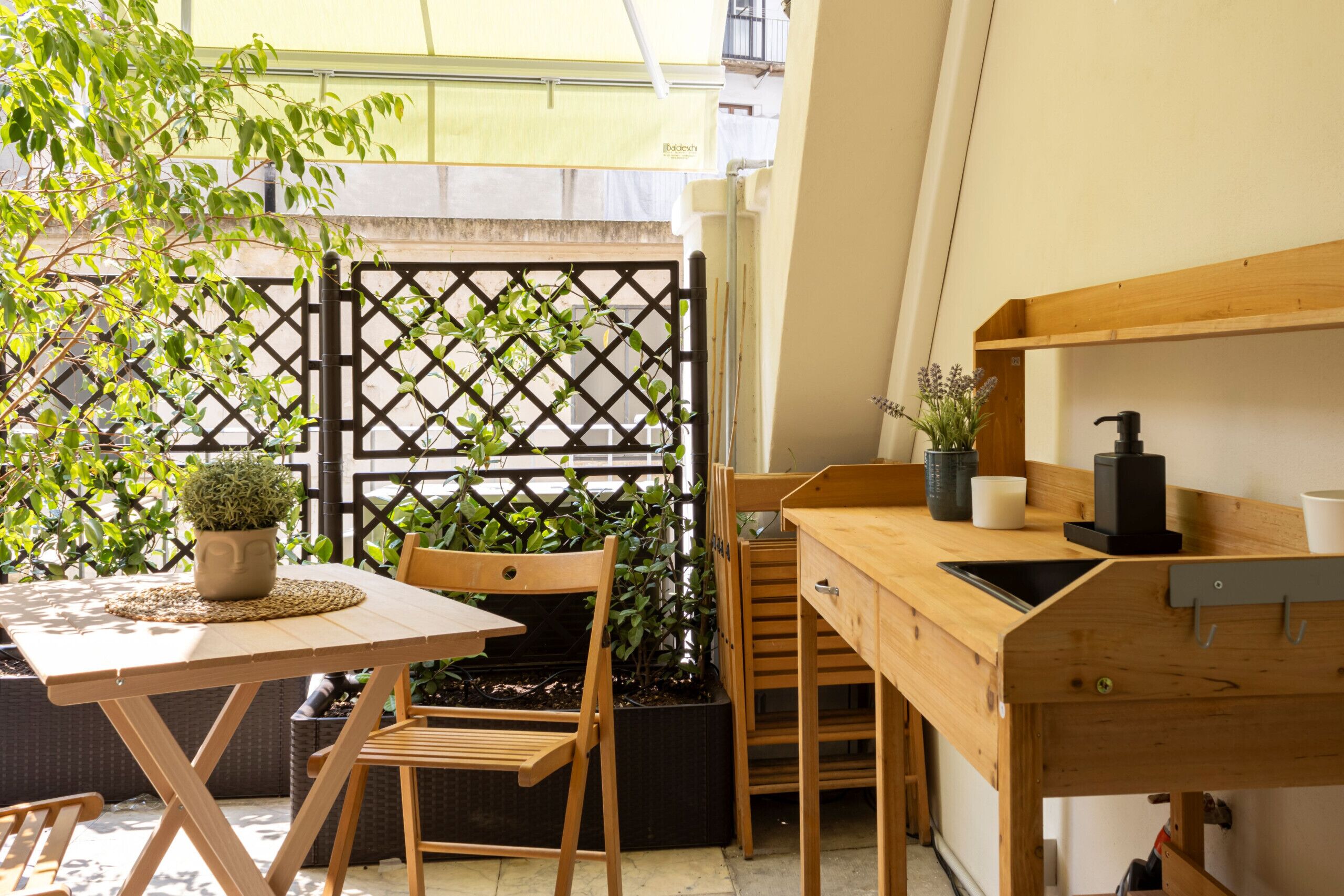 A compact kitchen area with a wooden table, chairs, and a sink. There is a potted plant on the table and a lattice fence in the background.