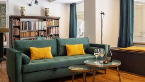 A green sofa with yellow cushions, a small round table with wine glasses and a bottle, and a bookshelf filled with books. - Turin Bed Books by Wonderful Italy (Turin)